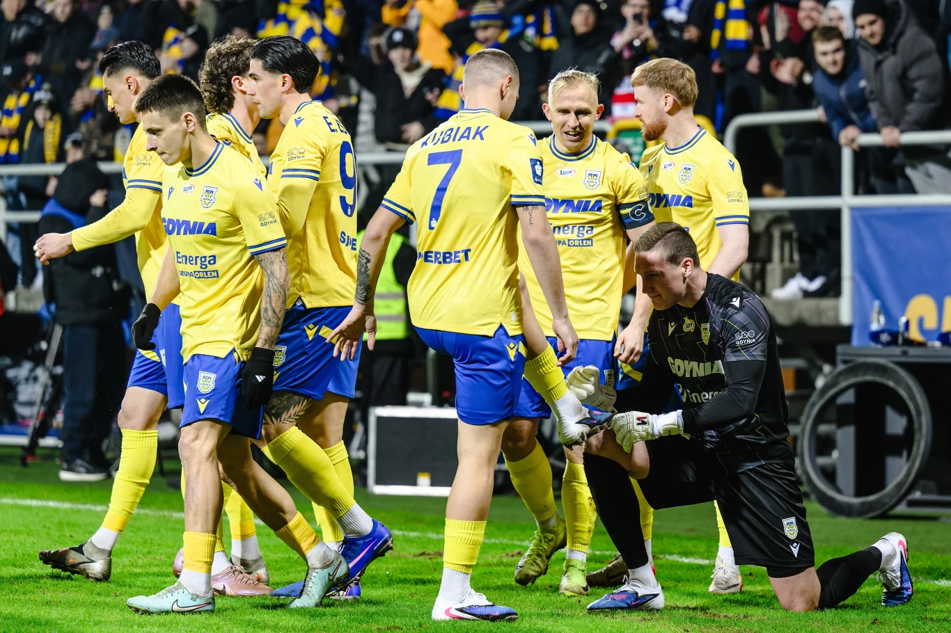 Piłkarze drużyny w żółto-niebieskich strojach świętują na murawie, jeden z zawodników w czarnym stroju klęczy i wykonuje gest triumfu, wokół niego stoją współtowarzysze z uśmiechami i zadowoleniem, w tle tłum kibiców ubranych w klubowe barwy.