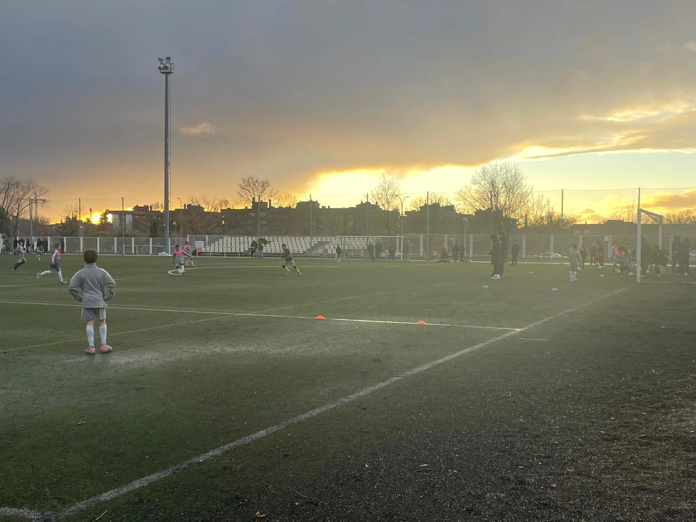 Dzieci grające w piłkę nożną na boisku podczas treningu, trener nadzorujący ćwiczenia, zachodzące słońce w tle nadaje scenie ciepłe oświetlenie, wokół boiska widoczne są pachołki wyznaczające strefy treningowe.