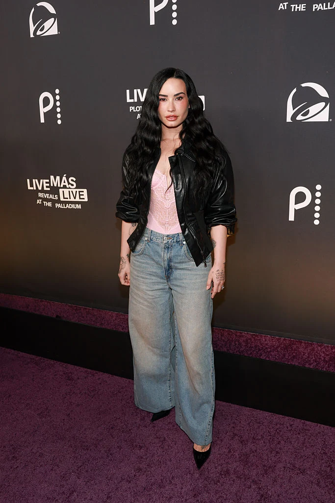 Kobieta o długich, ciemnych falowanych włosach pozuje na tle czarnej ścianki z logotypami, ubrana w czarną skórzaną kurtkę, jasnoróżowy top i szerokie niebieskie dżinsy oraz czarne buty na obcasie.