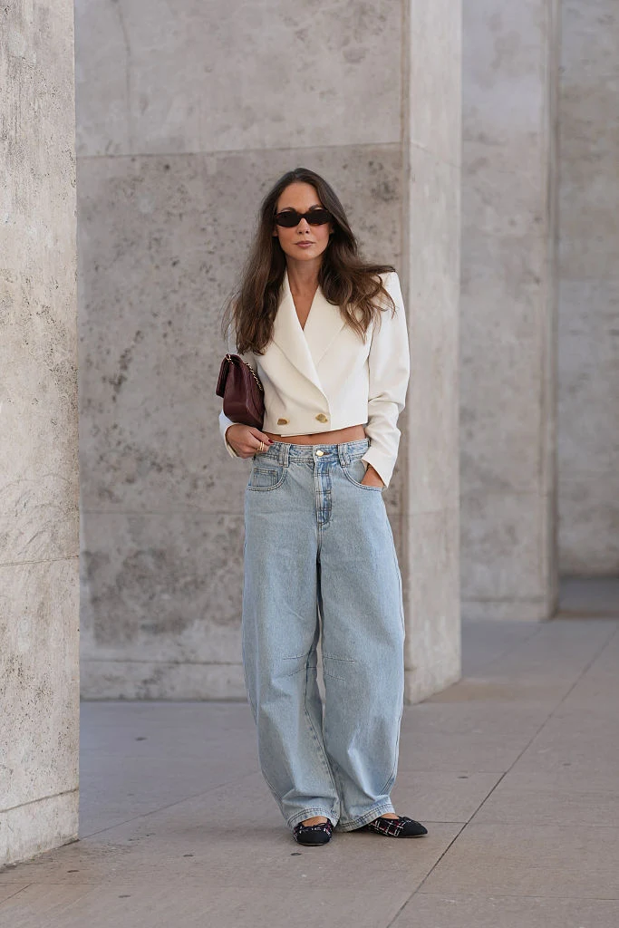 Kobieta ubrana w biały, krótki żakiet i szerokie, jasnoniebieskie dżinsy, trzymająca bordową kopertówkę i nosząca ciemne okulary przeciwsłoneczne, stoi przy betonowej ścianie.