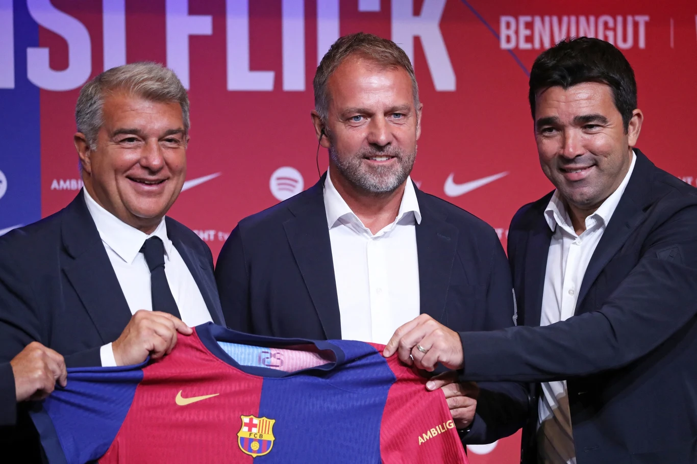 Laporta, Flick, Deco Laporta, Flick i Deco z koszulką FC Barcelony na tle ściany z napisem 'BENVINGUT' podczas prezentacji.