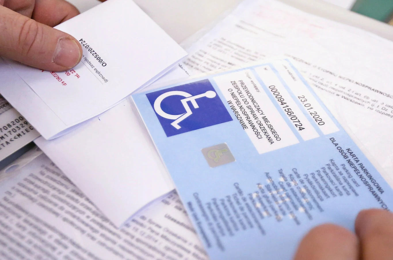 Karty parkingowe dla niepełnosprawnych się kończą. Problem w wielu powiatach. Karta parkingowa dla osoby niepełnosprawnej na tle dokumentów urzędowych, widoczny symbol wózka inwalidzkiego