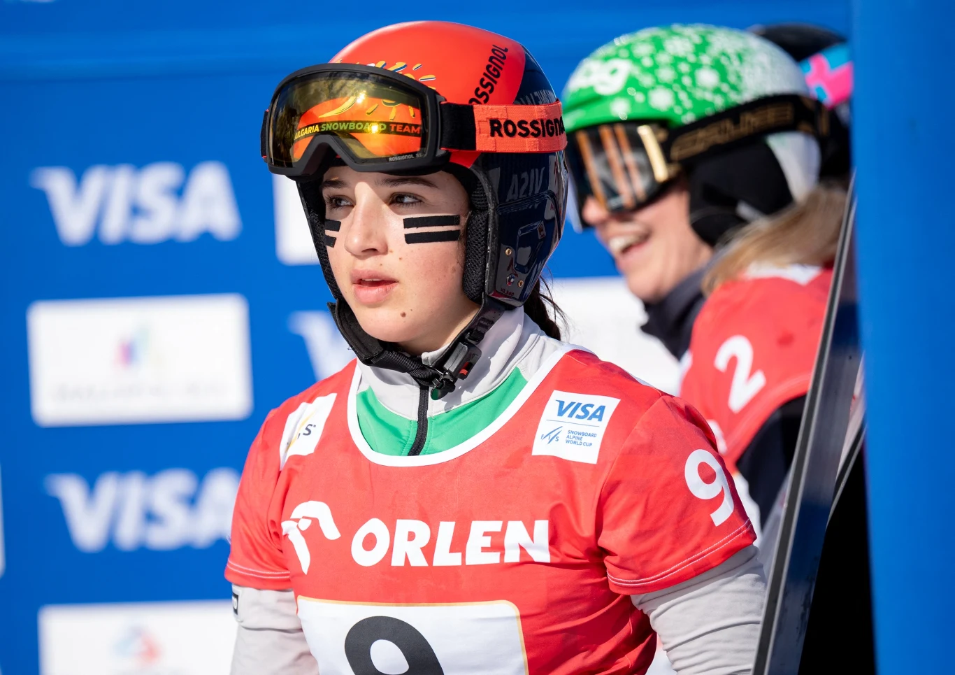 Zawodniczka ubrana w czerwony numer startowy z logo Orlen i wizjerem narciarskim przygotowuje się do wyścigu na tle banerów sponsorskich, obok niej inna uczestniczka w zielonym kasku i okularach."