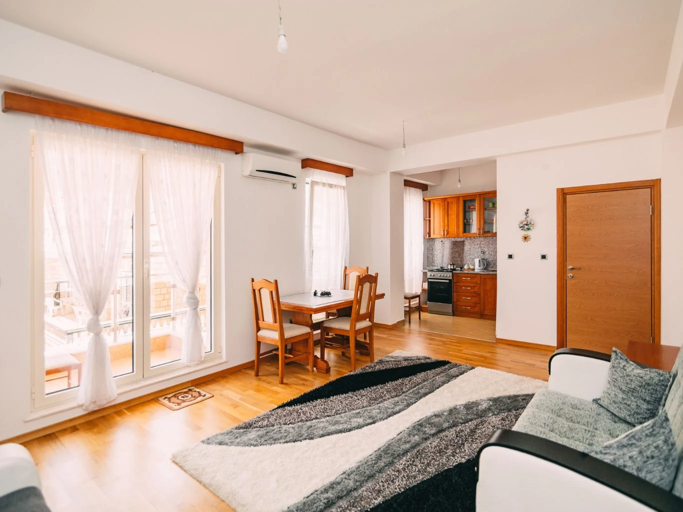 Uniwersalny i bardzo modny kolor do salonu i kuchni. Jasny salon z drewnianym stołem i sofą, płynnie przechodzący w kuchnię z jasnymi szafkami i kuchenką.