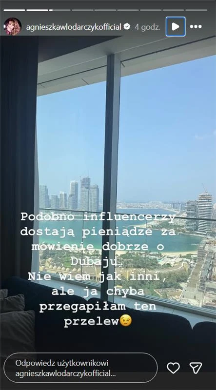 Panorama Dubaju z wysokiego piętra budynku, duże okna ukazujące nowoczesne wieżowce, fragment wybrzeża i błękitne niebo; na pierwszym planie widoczna część ciemnej kanapy oraz zasłona, na zdjęciu naniesiony tekst w języku polskim.