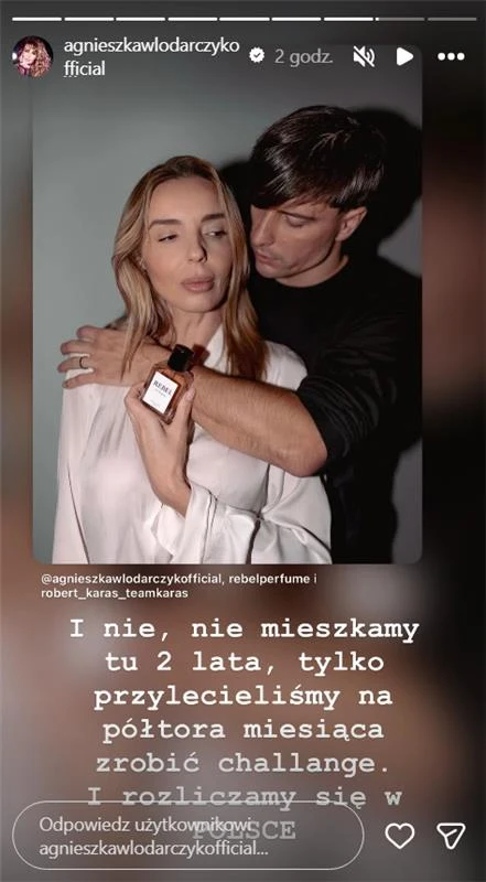 Kobieta w białej koszuli trzymająca perfumy w ręku, obejmowana przez mężczyznę stojącego za nią; oboje mają spokojne, poważne wyrazy twarzy, tło neutralne.