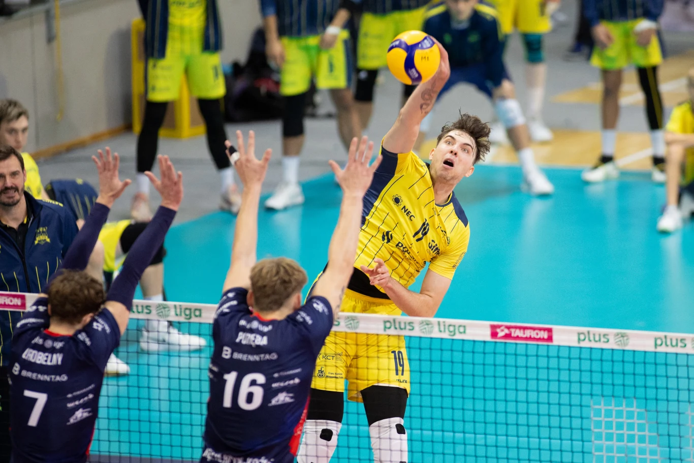 Siatkarz w żółtym stroju oddaje dynamiczny atak nad siatką, podczas gdy dwóch zawodników w granatowych koszulkach próbuje go zablokować, kibice oraz drużyna w tle obserwują akcję.
