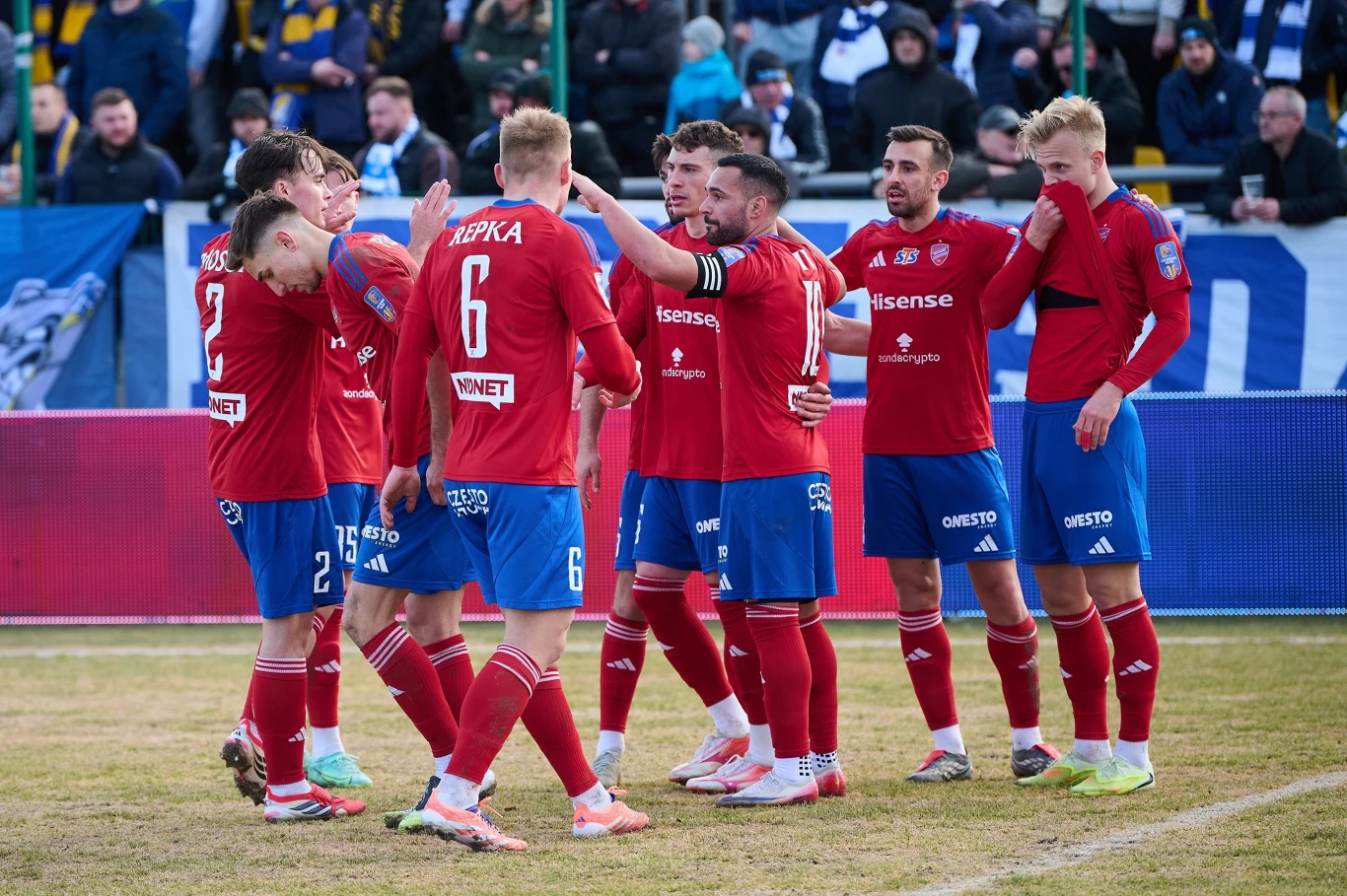 Grupa piłkarzy w czerwonych koszulkach świętuje zdobycie bramki, stojąc razem na murawie, otoczona trybunami z kibicami i niebiesko-białymi flagami w tle.