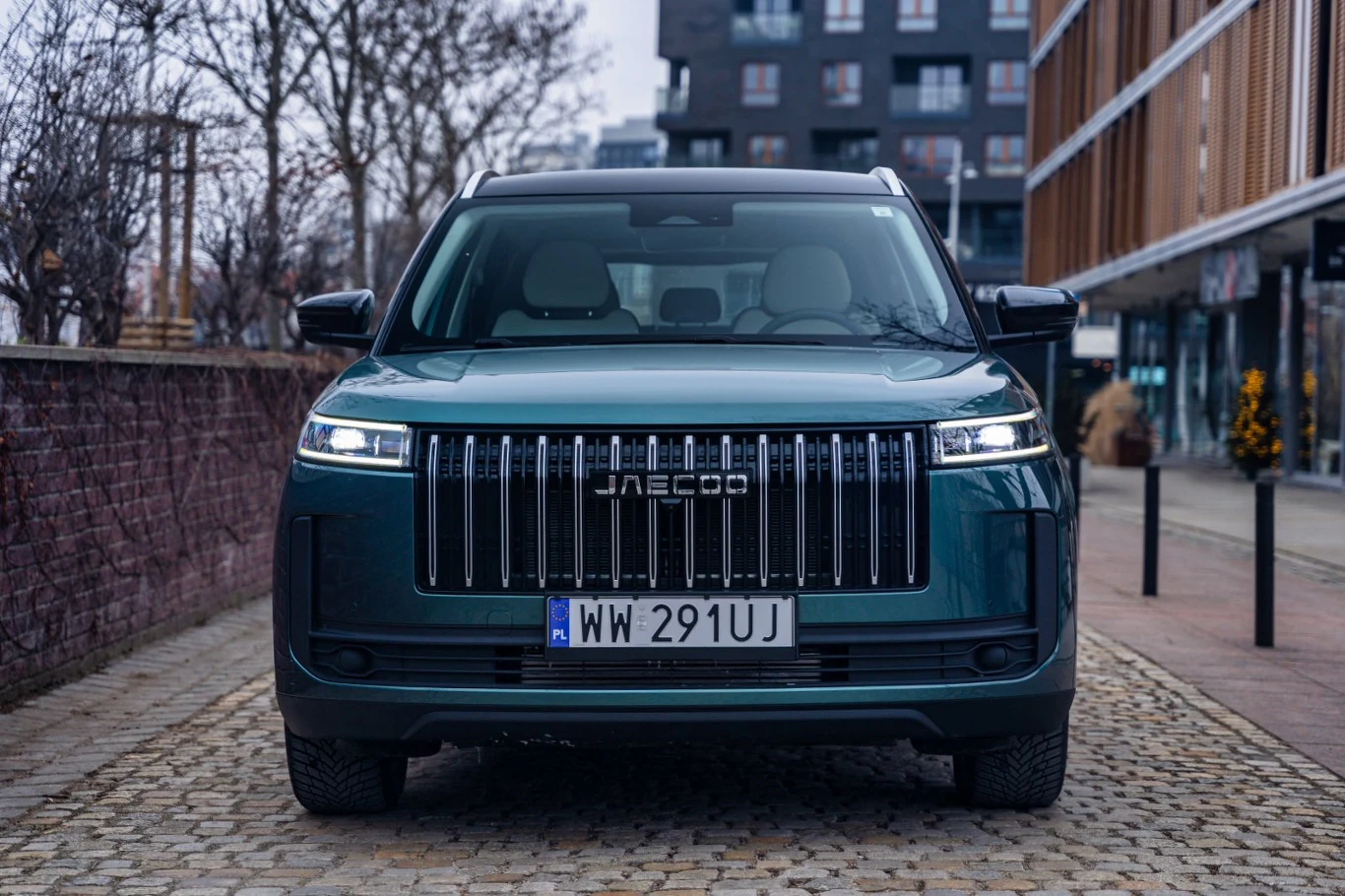 Duży, nowoczesny SUV marki Jaecoo o ciemnozielonym kolorze, z charakterystycznym pionowym grillem i prostokątnymi reflektorami, zaparkowany na brukowanej ulicy w miejskiej przestrzeni.