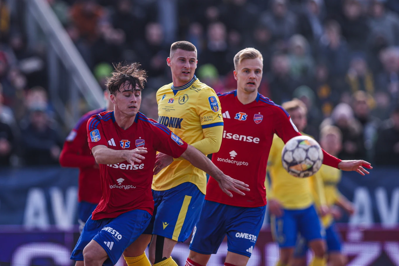 Trzech piłkarzy w trakcie meczu walczy o piłkę, zawodnicy w czerwonych i żółtych strojach rywalizują ze sobą na tle rozmytej publiczności na trybunach.