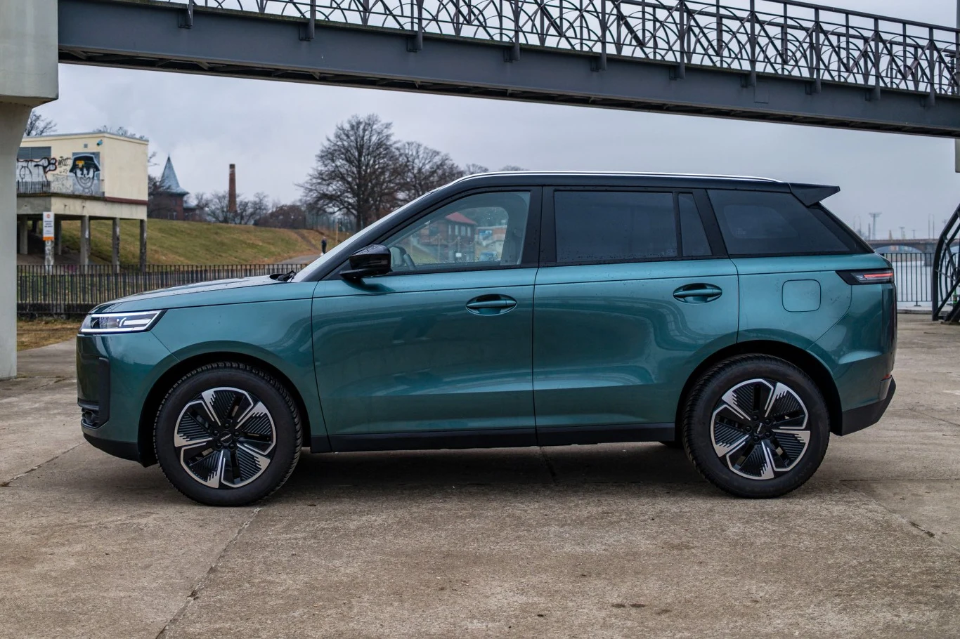 Nowoczesny, zielony SUV o aerodynamicznej sylwetce, ciemnych szybach oraz stylowych felgach stoi na betonowej nawierzchni, w tle widoczny metalowy most oraz drzewa bez liści.