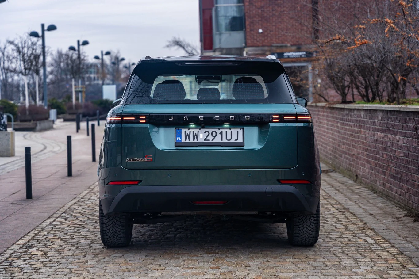 Ciemnozielony SUV marki Jaecoo z jasno oznaczoną rejestracją, ustawiony tyłem na brukowanej ulicy, otoczony niskim murem z cegły i miejską przestrzenią z latarniami oraz nagimi drzewami w tle.