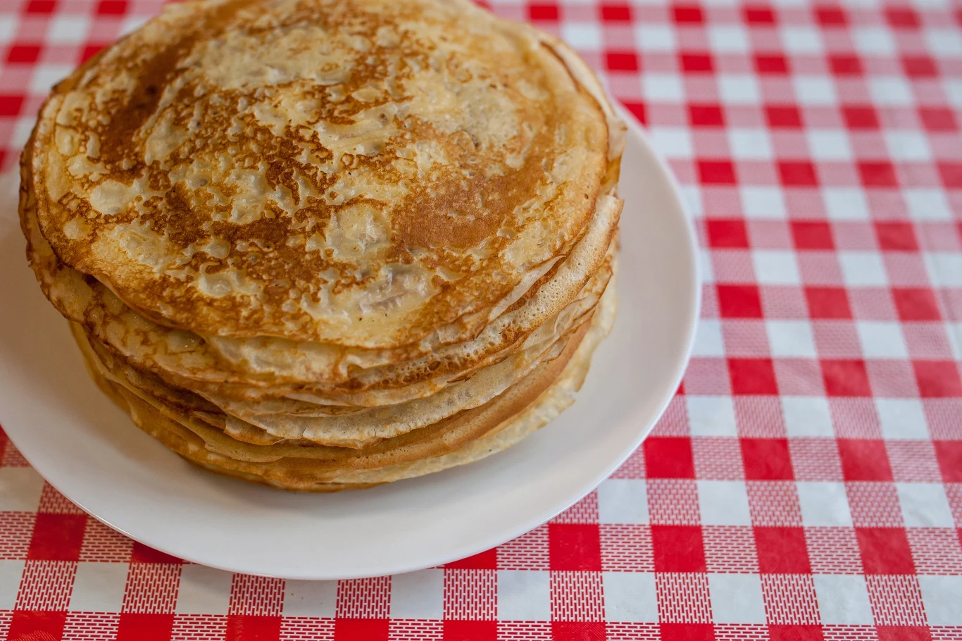 Stos naleśników ułożony na białym talerzu, ustawionym na czerwono-białym obrusie w kratę.
