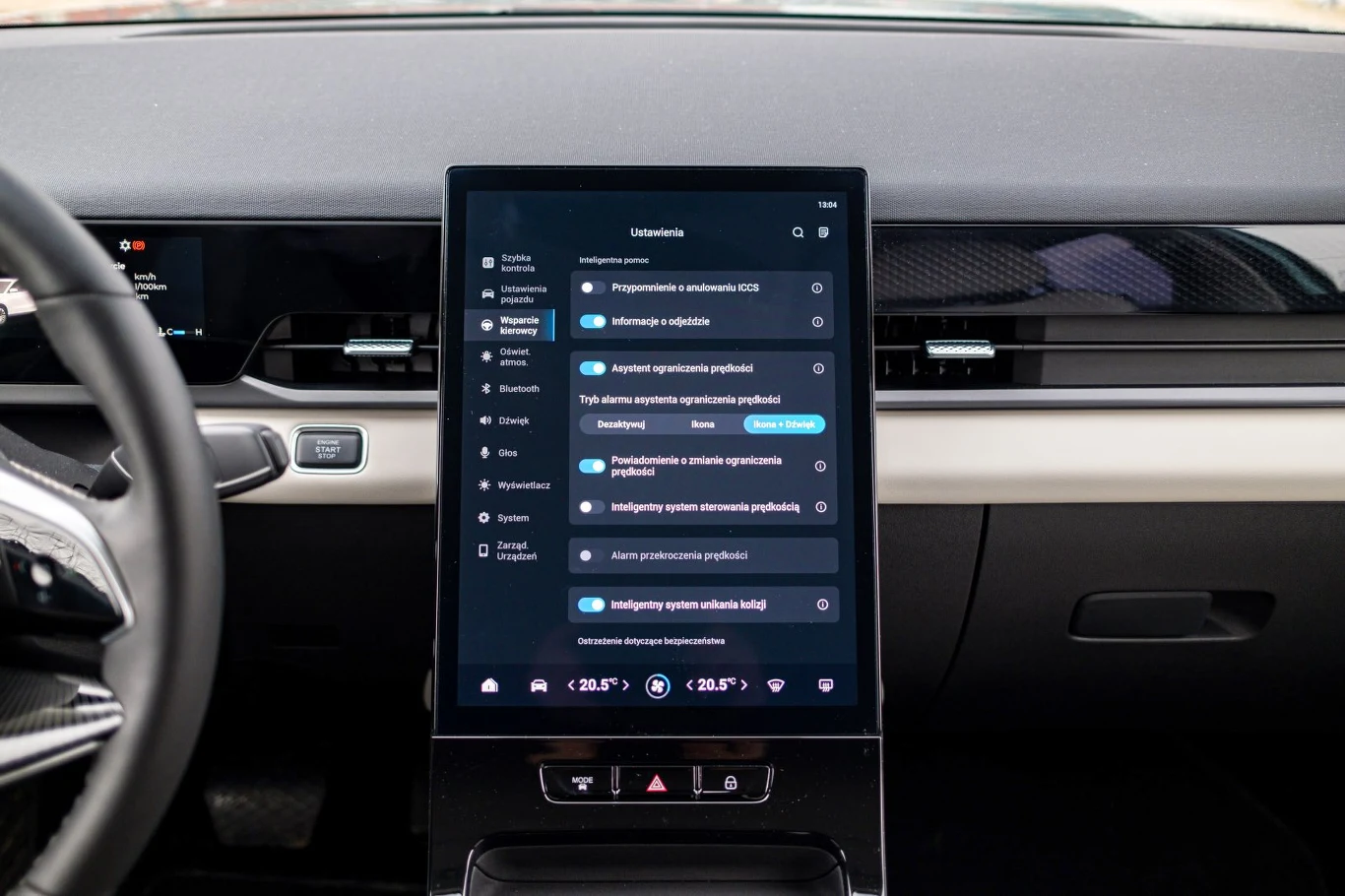 Wnętrze nowoczesnego samochodu z dużym, pionowym ekranem dotykowym na konsoli centralnej, wyświetlającym ustawienia systemowe oraz opcje zarządzania pojazdem, otoczone minimalistyczną deską rozdzielczą i elementami kierownicy po lewej stronie.