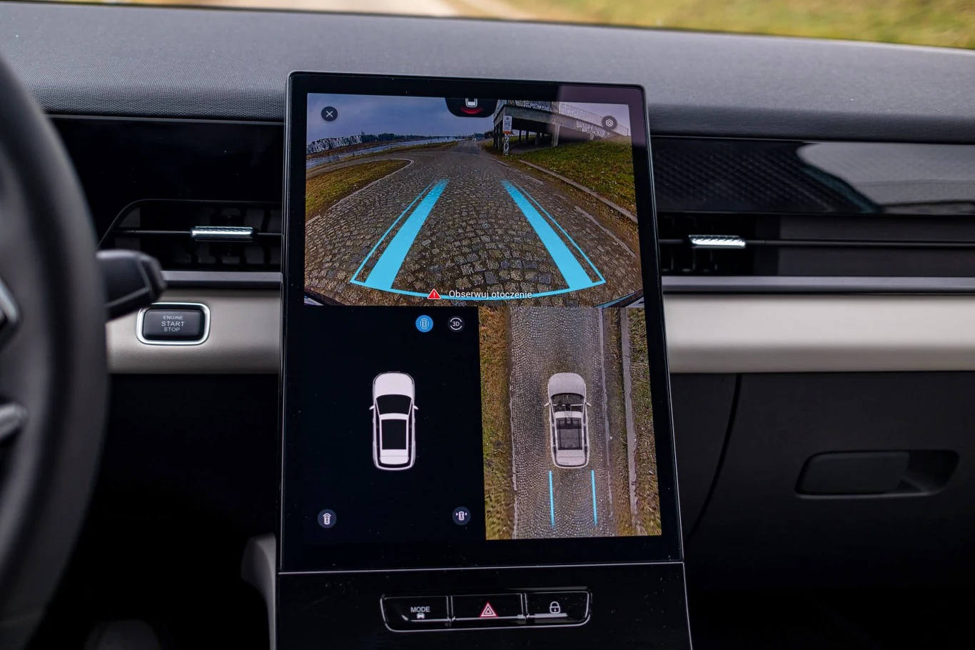 Nowoczesny ekran dotykowy w samochodzie prezentujący widok z kamery cofania oraz system asystenta parkowania z wizualizacją toru jazdy i rozmieszczeniem pojazdu na drodze.