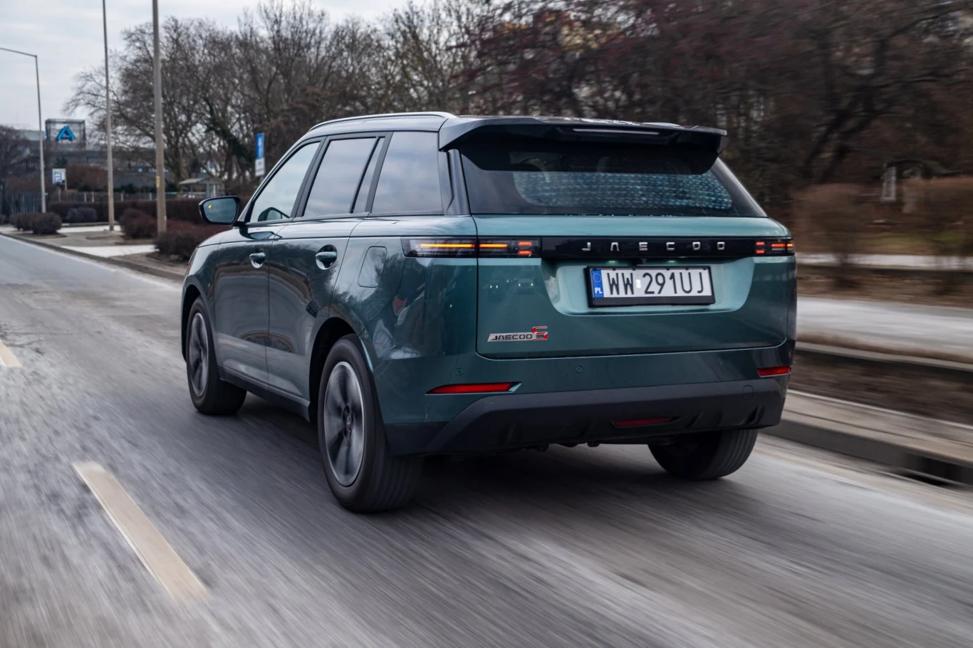 Jadeozielony SUV marki Jaecoo jedzie po pustej drodze miejskiej, otoczony bezlistnymi drzewami, widoczna jest rejestracja i nowoczesny design tylnej części pojazdu.