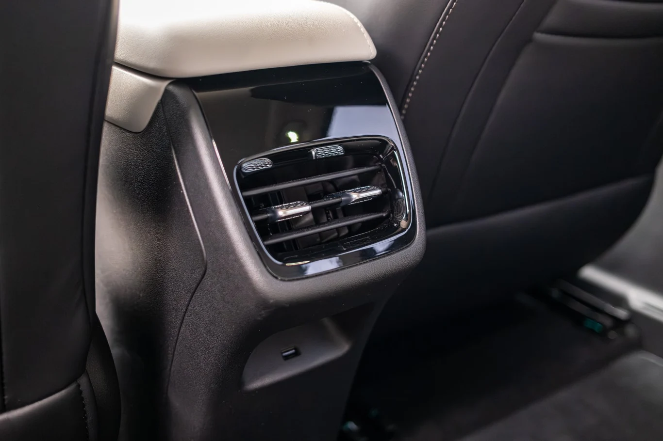 Czarny panel z kratką nawiewu powietrza zamontowany na tylnej części środkowego tunelu w samochodzie, otoczony czarną tapicerką z białym przeszyciem oraz jasnym elementem obiciowym na górze.
