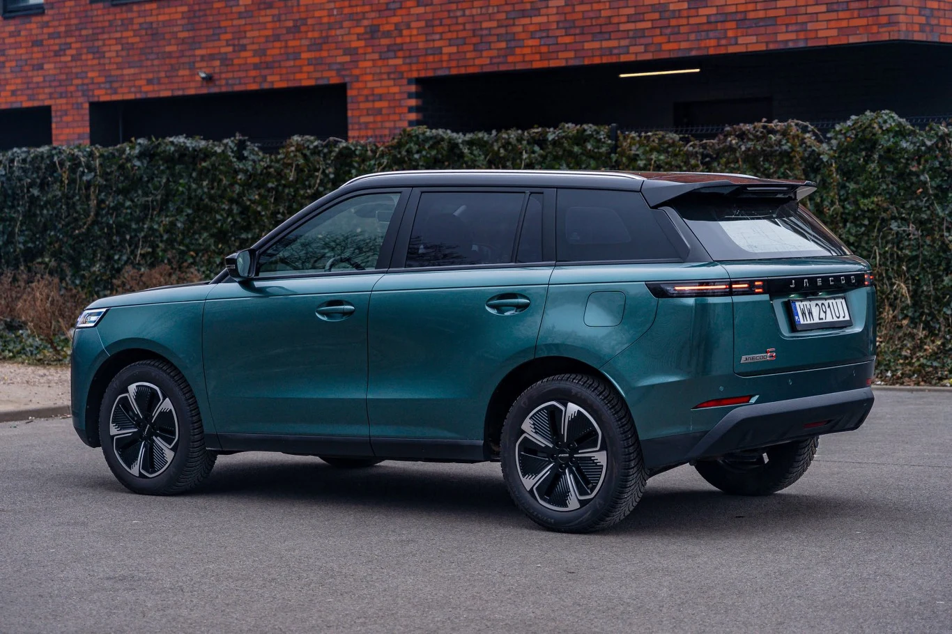 Zielony SUV o nowoczesnym, opływowym kształcie z przyciemnianymi szybami i czarnymi felgami, zaparkowany na tle zielonego żywopłotu i ceglanego budynku.