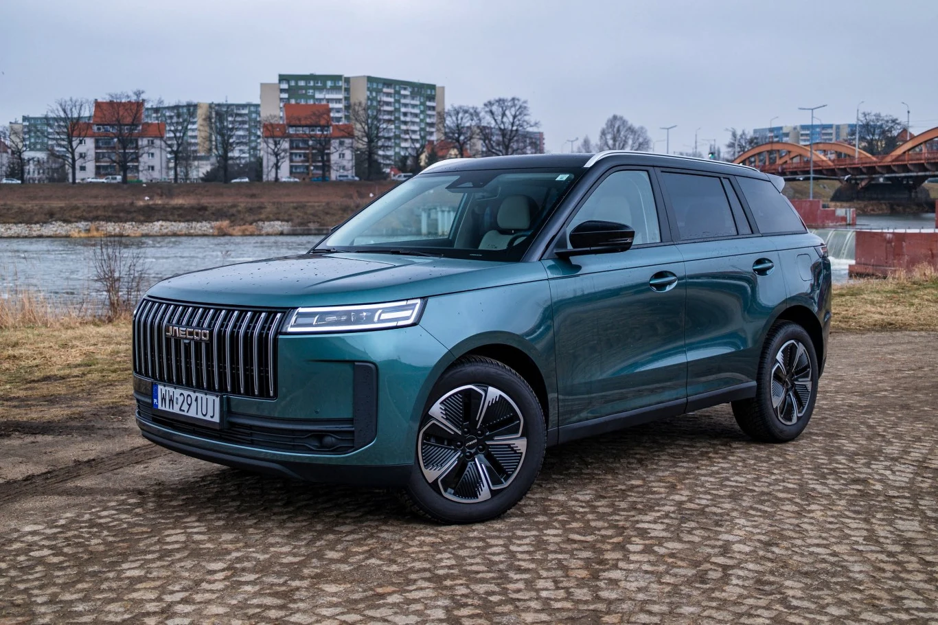 Zielony, nowoczesny SUV z czarnymi felgami stoi na brukowanej nawierzchni, w tle widoczna rzeka oraz zabudowa mieszkaniowa z blokami i mostem.