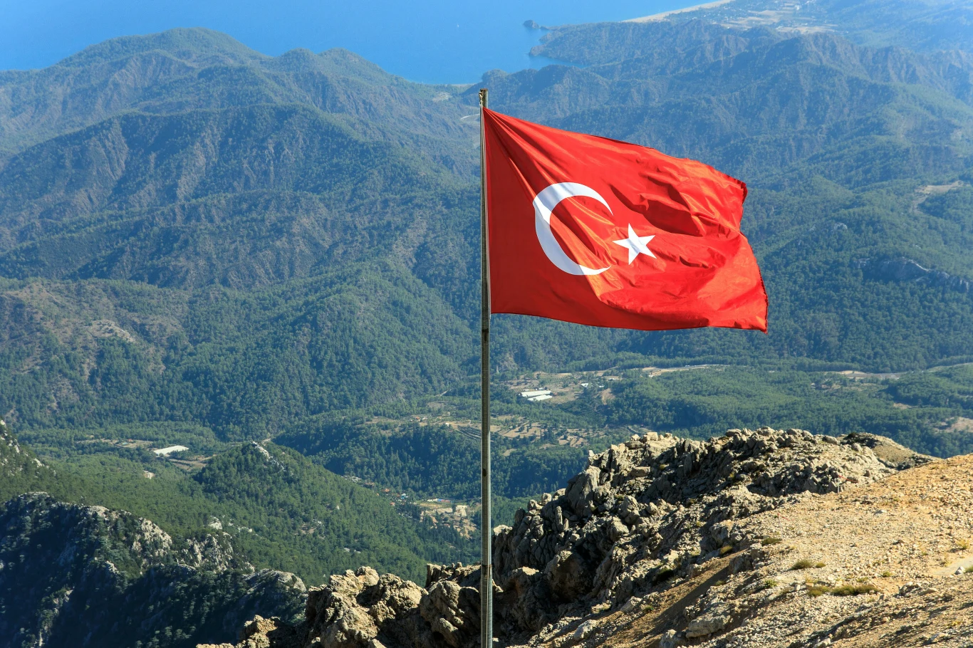 MSZ dodał na listę ostrzeżeń Adanę w Turcji. Zdjęcie ilustracyjne Czerwona flaga Turcji z półksiężycem i gwiazdą na tle gór, lasu i wybrzeża z morzem w oddali.