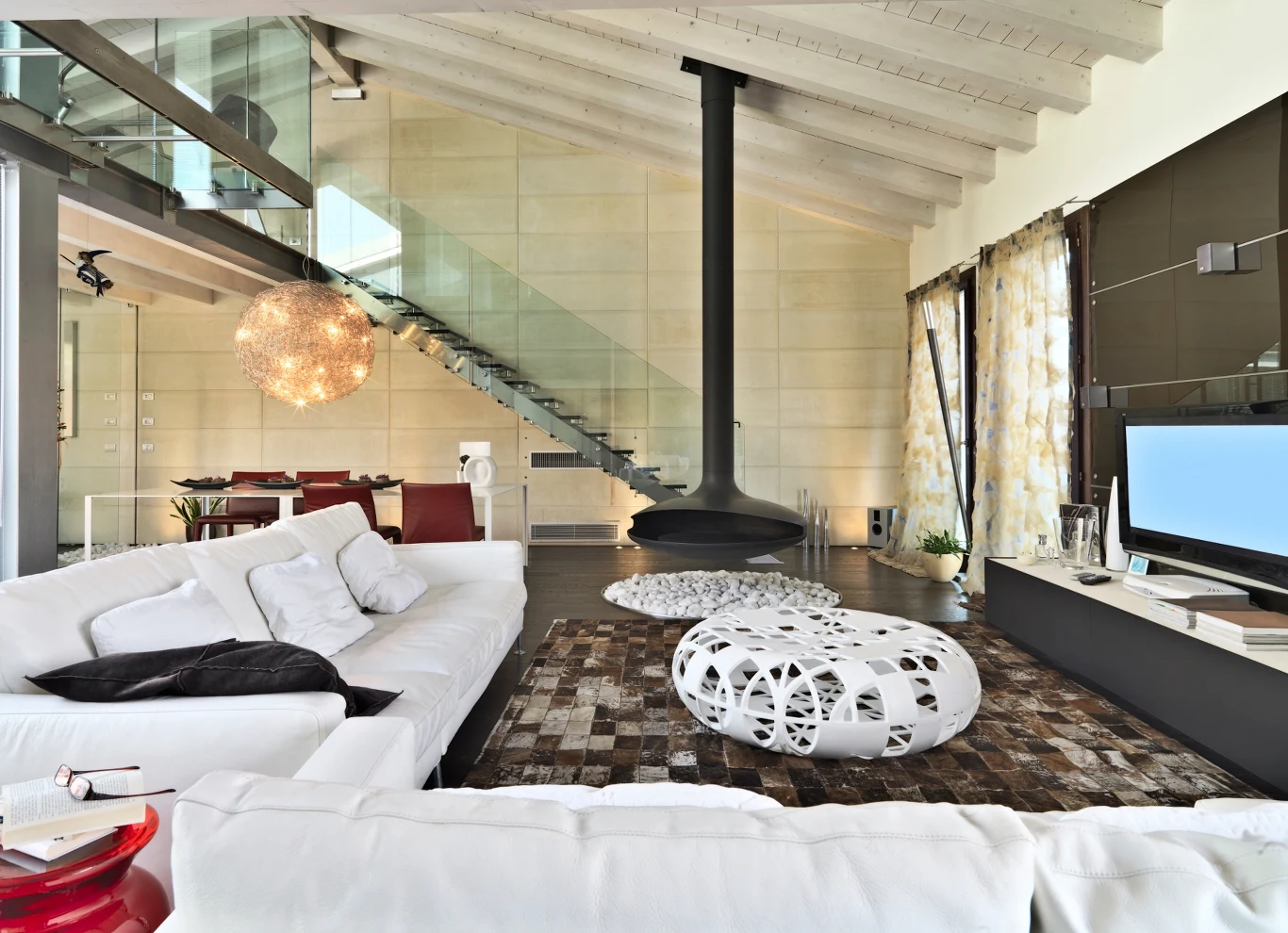 Przestronny i nowoczesny salon z białymi sofami, wiszącym kominkiem, designerskim stolikiem o ażurowej strukturze, szklaną klatką schodową oraz dużym telewizorem naprzeciwko wygodnej strefy wypoczynkowej.