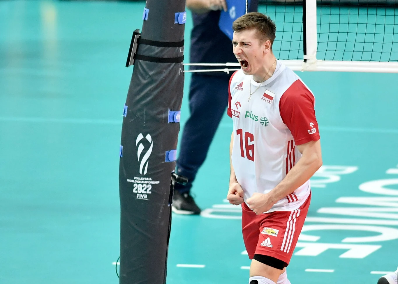 Siatkarz w biało-czerwonym stroju z numerem 16 wyraża silne emocje, zaciskając pięści i krzycząc, stojąc blisko siatki na boisku podczas meczu siatkówki.