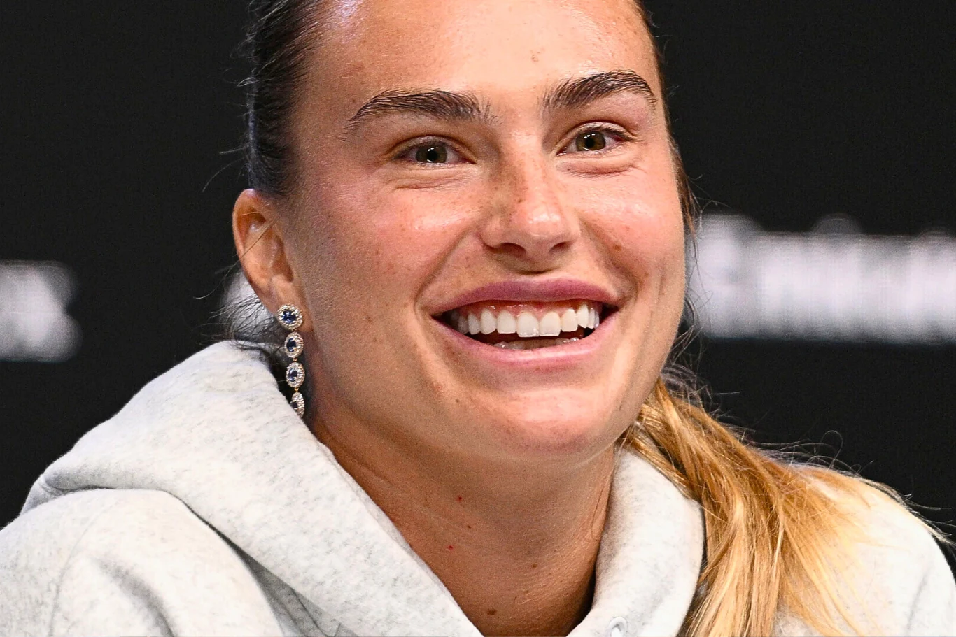 Aryna Sabalenka Aryna Sabalenka siedzi na ciemnym tle, ubrana w szarą bluzę z kapturem, blond włosy związane w kucyk.