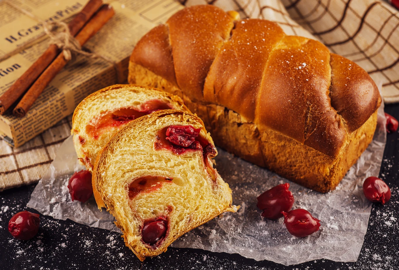 Maślana chałka z owocowym nadzieniem wiśniowym, podana na pergaminie, z rozrzuconymi wokół świeżymi wiśniami oraz laskami cynamonu w tle.