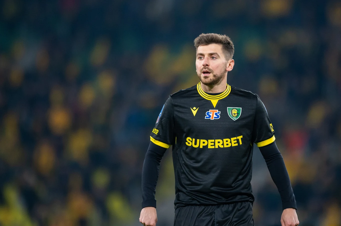 Piłkarz w czarnym stroju GKS Katowice z logo sponsora SUPERBET stoi na boisku podczas meczu, skupiony, z lekko otwartymi ustami, na tle trybun wypełnionych rozmytymi kibicami.