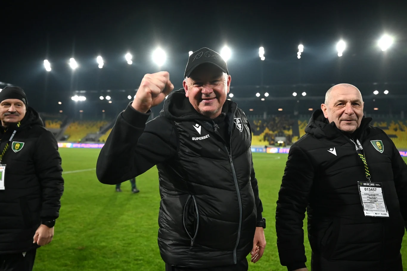 Trener drużyny piłkarskiej w czarnej kurtce i czapce zaciśniętą pięścią okazuje radość i triumf na boisku, pośród towarzyszących mu członków sztabu, w tle jasno oświetlone trybuny stadionu.