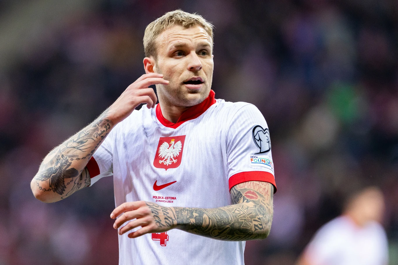 Piłkarz w koszulce reprezentacji Polski z widocznym orłem na piersi, licznymi tatuażami na ramionach i rękach, stoi na murawie w trakcie meczu, skupiony na grze.