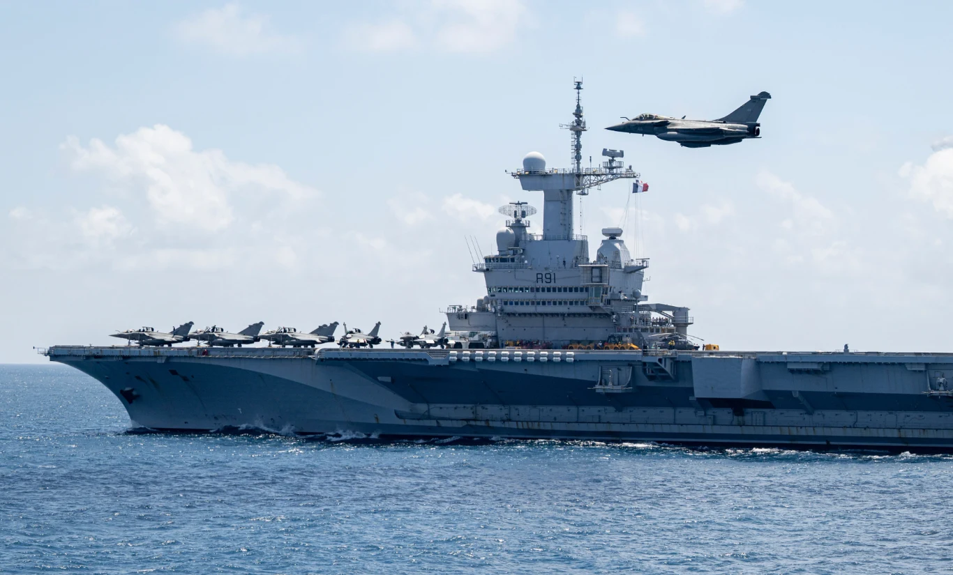 Lotniskowiec płynący po morzu z myśliwcami ustawionymi na pokładzie, nad okrętem przelatuje odrzutowiec wojskowy; nad mostkiem widoczna francuska flaga.