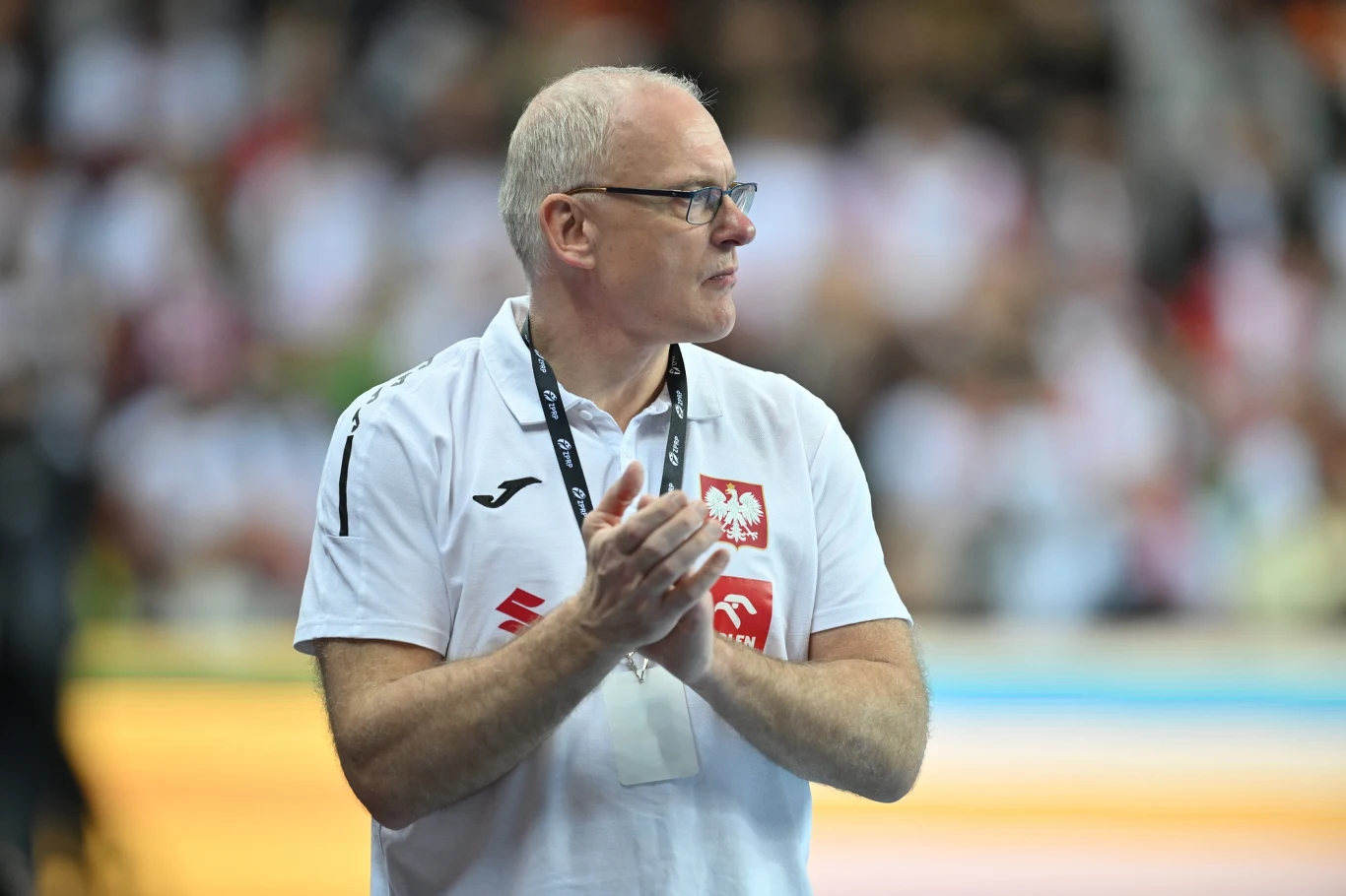 Mężczyzna w okularach o siwych włosach ubrany w białą koszulkę z herbem Polski i symbolami sportowymi, stoi na tle rozmytej publiczności, trzymając dłonie złożone jak do oklasków.