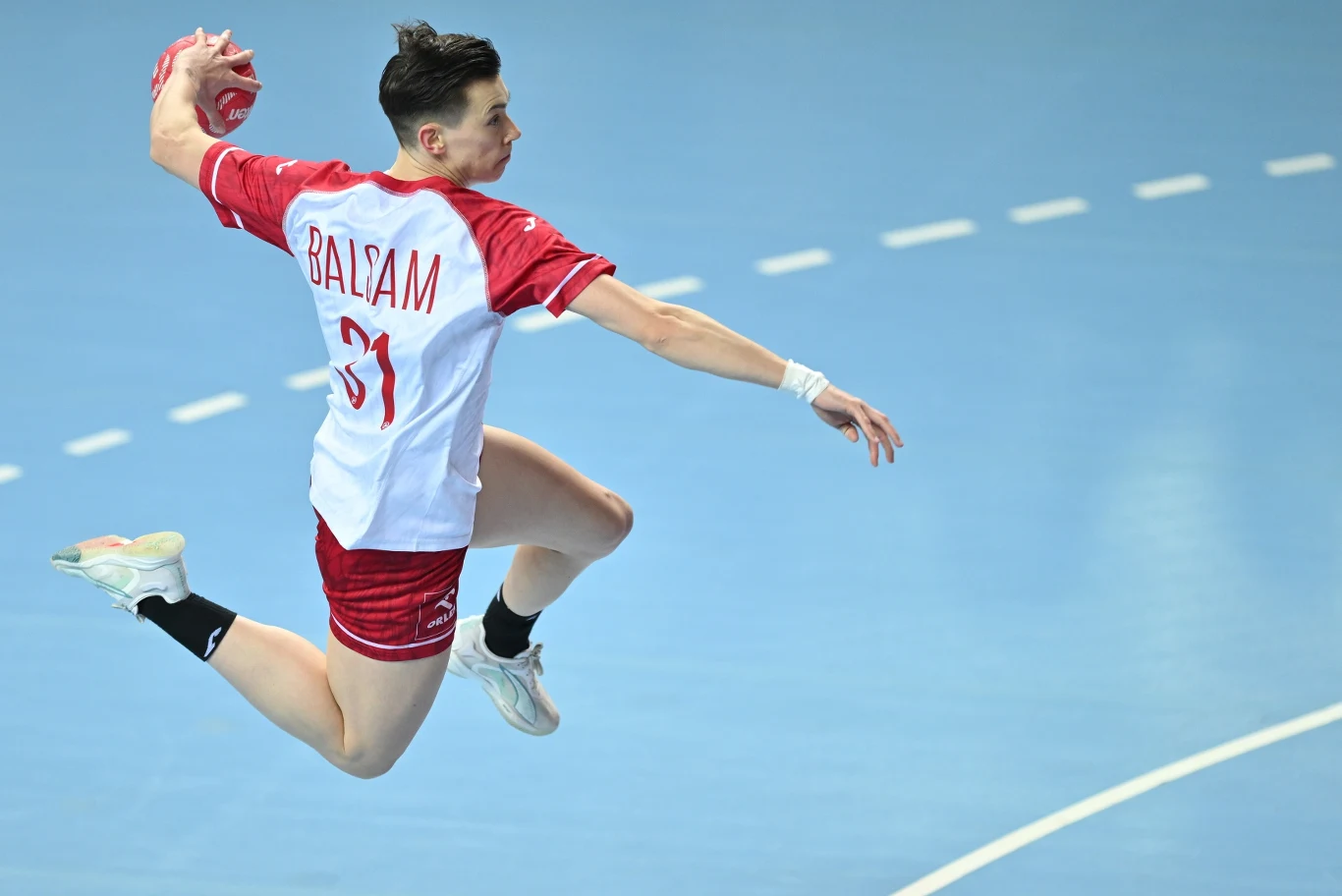 Zawodnik w czerwono-białym stroju sportowym podczas dynamicznego wyskoku, z piłką ręczną trzymaną w prawej dłoni, w trakcie akcji na niebieskim parkiecie hali sportowej.