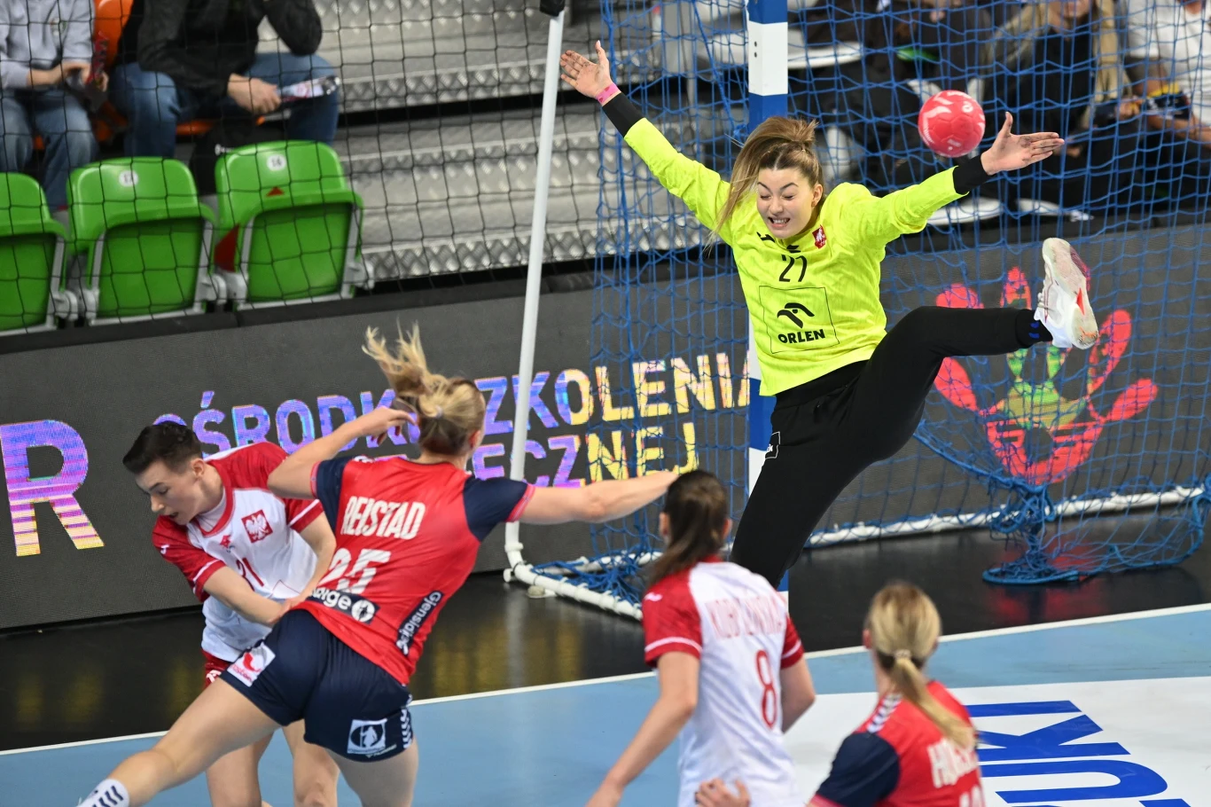 Bramkarka w jaskrawożółtym stroju wyskakuje w powietrze z szeroko rozłożonymi ramionami i nogami, próbując obronić rzut na bramkę, podczas gdy zawodniczki w czerwono-białych oraz czerwono-granatowych strojach walczą o piłkę przy bramce na tle trybun z ...