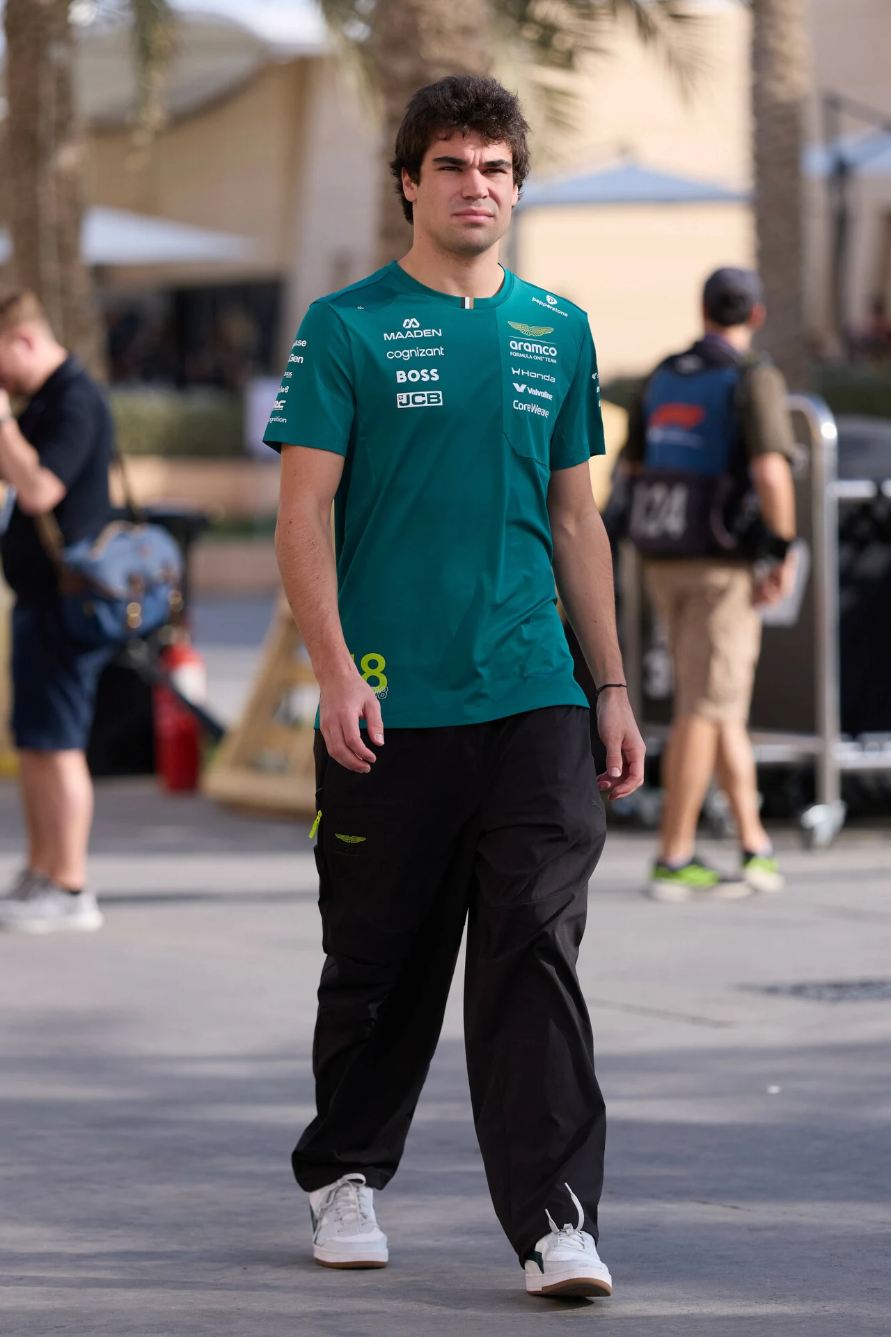 Mężczyzna w zielonej koszulce zespołu wyścigowego Aston Martin i czarnych spodniach spaceruje chodnikiem, w tle widoczni są inni ludzie oraz palmy i zabudowania toru wyścigowego.