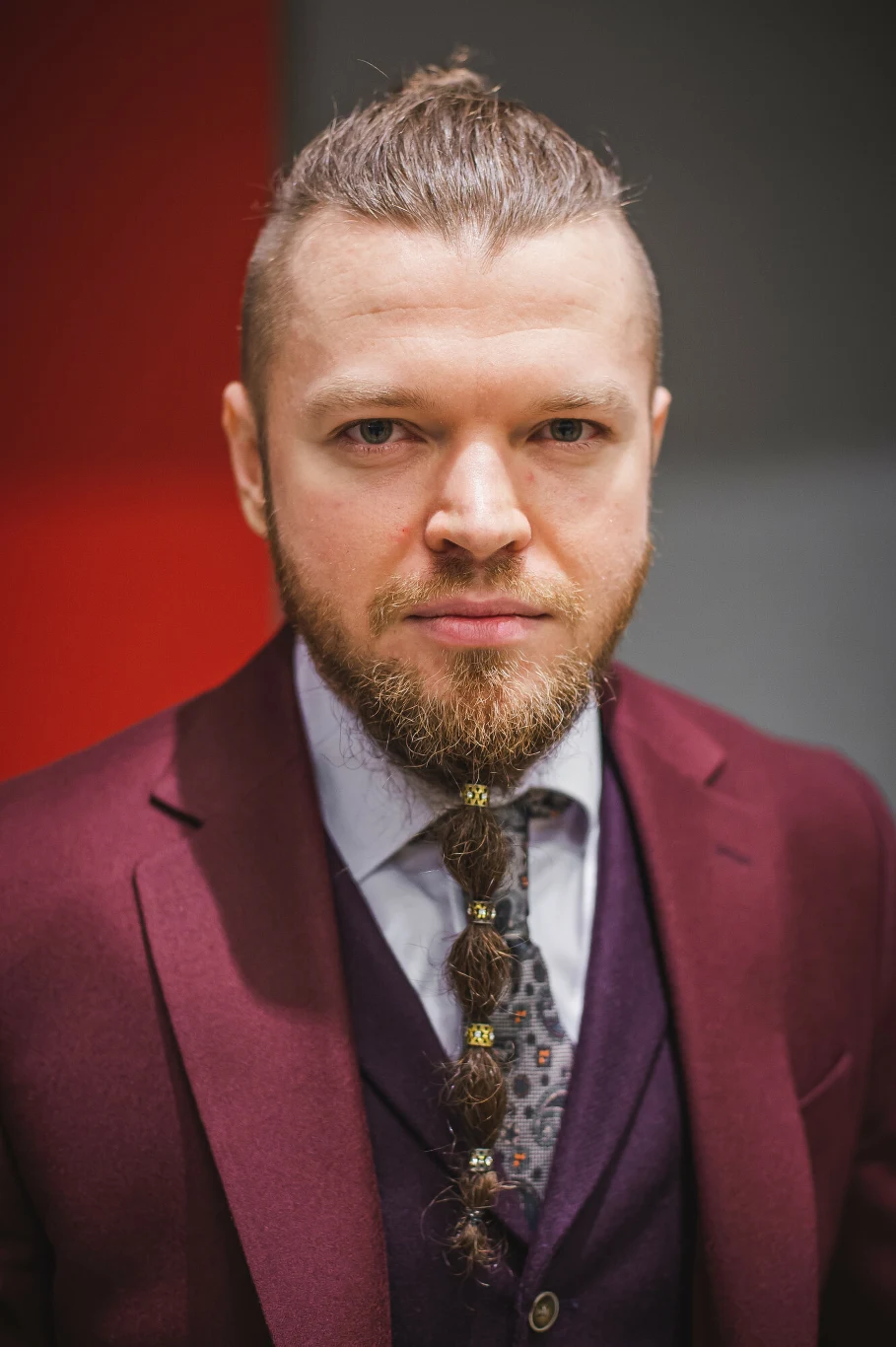 Mężczyzna z brodą zaplecioną w warkocz i spiętymi gumkami, ubrany w bordowy garnitur, fioletową kamizelkę, jasną koszulę oraz krawat w delikatny wzór, patrzący prosto w obiektyw na tle czerwono-szarej ściany.