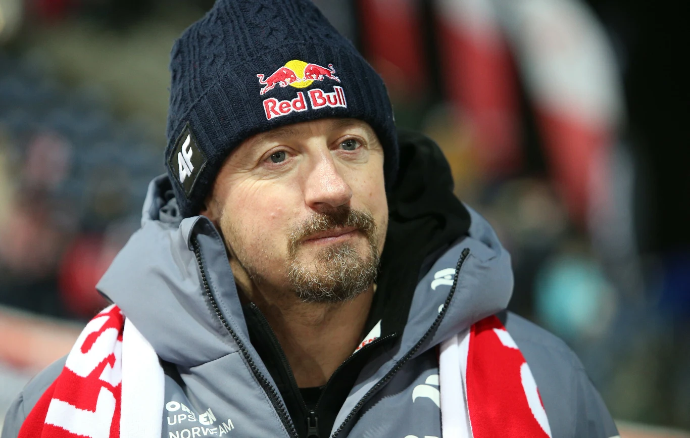 Mężczyzna w ciemnej zimowej czapce z logo Red Bull oraz w kurtce i szaliku sportowym, spoglądający spokojnie przed siebie na tle rozmytej sceny stadionowej.