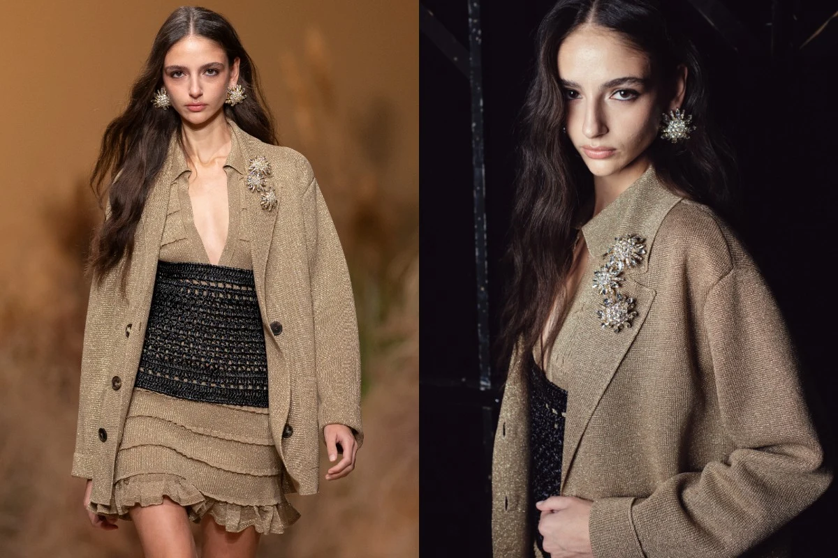 Młoda kobieta w beżowej marynarce z ozdobną broszką nosi krótką, falbaniastą sukienkę w odcieniach beżu oraz czarny gorset z siateczkowej tkaniny, a jej stylizację dopełniają duże, dekoracyjne kolczyki.