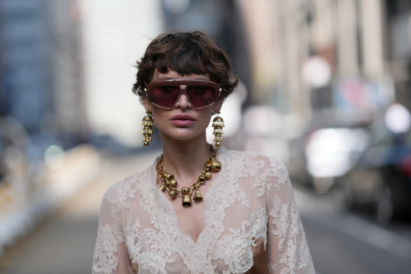 Stylowa kobieta z krótką fryzurą i dużymi okularami przeciwsłonecznymi, ubrana w koronkową bluzkę w jasnym kolorze, z okazałymi złotymi kolczykami i naszyjnikiem, stoi na ulicy miasta w tle, skoncentrowana na modnych akcesoriach.