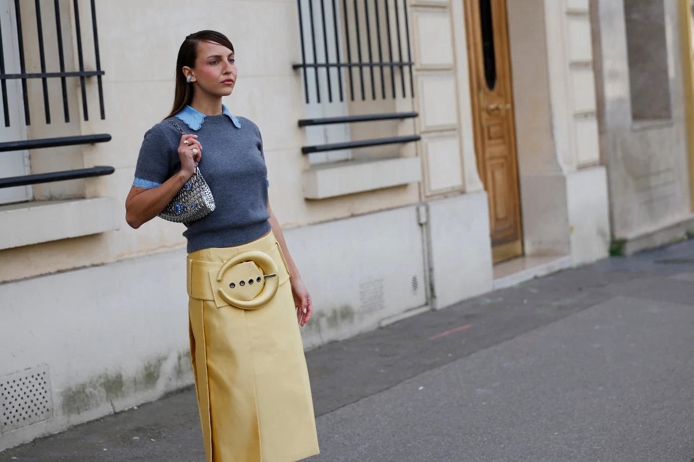 Kobieta w grafitowym swetrze z jasnoniebieskim kołnierzykiem i żółtej spódnicy z dużą dekoracyjną aplikacją w kształcie guzika, trzymająca srebrną torebkę, spaceruje po miejskim chodniku na tle jasnej elewacji budynku z charakterystycznymi oknami zabez...