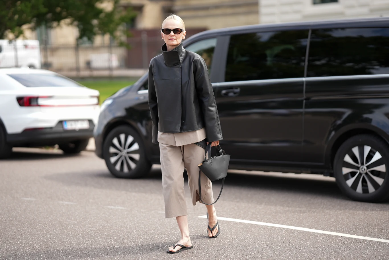 Elegancka kobieta o krótkich, siwych włosach ubrana w czarną kurtkę, szare spodnie 3/4 i klapki, trzymająca czarną torebkę i nosząca czarne okulary przeciwsłoneczne, idzie ulicą obok czarnego samochodu.