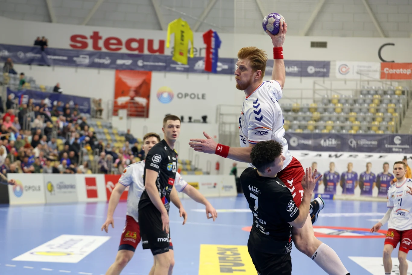 Zawodnik w białej koszulce wyskakuje z piłką do rzutu na bramkę, trzymany przez przeciwnika w czarnym stroju, podczas dynamicznej akcji meczu piłki ręcznej, na tle trybun z kibicami i banerami reklamowymi.