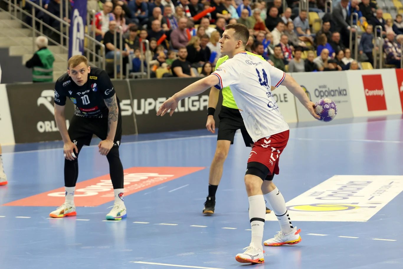 Zawodnik w białej koszulce i czerwonych spodenkach przygotowuje się do rzutu podczas meczu piłki ręcznej, podczas gdy inny zawodnik w czarnym stroju obserwuje jego ruchy, a sędzia stoi w tle na niebieskim parkiecie w hali pełnej kibiców.