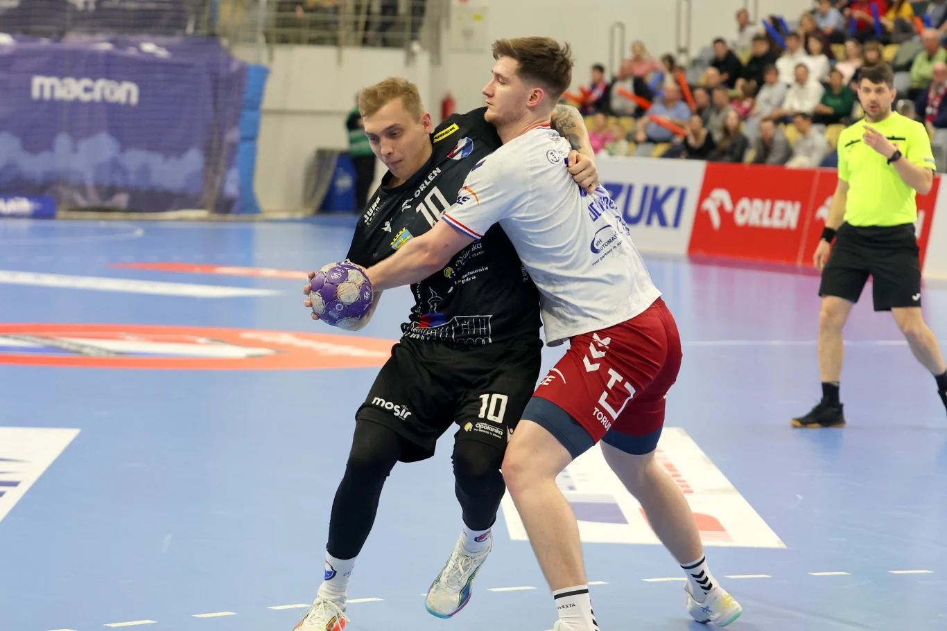 Dwóch szczypiornistów walczy o piłkę na hali sportowej podczas meczu, jeden ubrany na czarno z piłką w dłoni, drugi w białej koszulce i czerwonych spodenkach próbuje go zatrzymać, w tle widoczny sędzia oraz publiczność na trybunach.