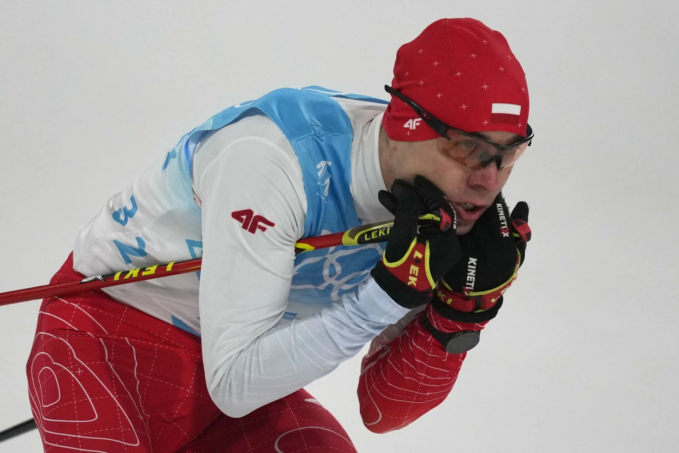 Narciarz biegowy w czerwonym stroju z białymi i niebieskimi elementami, pochylony do przodu w trakcie wyścigu, z kijami narciarskimi opartymi o policzki, na głowie czerwona czapka i sportowe okulary ochronne.