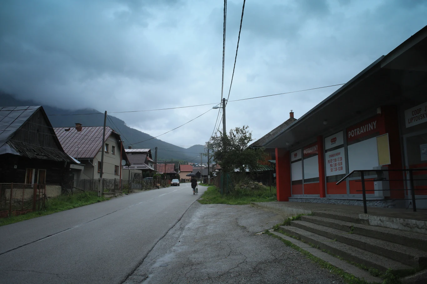 Liptovska Luzna, wieś położona w górach Nizne Tatry Opustoszała ulica w małym miasteczku, po lewej stronie stare, drewniane domy mieszkalne, po prawej stronie zamknięty sklep z czerwonymi oznaczeniami, na horyzoncie góry pod pochmurnym niebem, a pojedyncza osoba idzie chodnikiem w oddali.