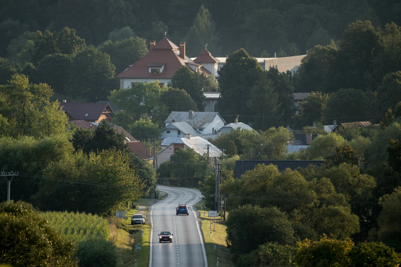 Zborov Kręta asfaltowa droga prowadzi przez zieloną wieś w kierunku zabytkowego kompleksu budynków z czerwonymi dachami, otoczonych gęstym lasem i drzewami, po drodze mijają się pojedyncze samochody.