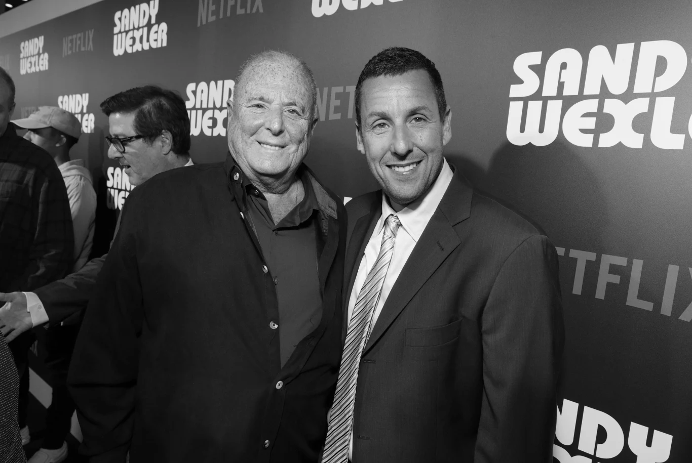 Dwaj uśmiechnięci mężczyźni stoją obok siebie na tle ścianki z napisem 'Sandy Wexler' i 'Netflix', ubrani w eleganckie marynarki, jeden z nich ma na sobie krawat.