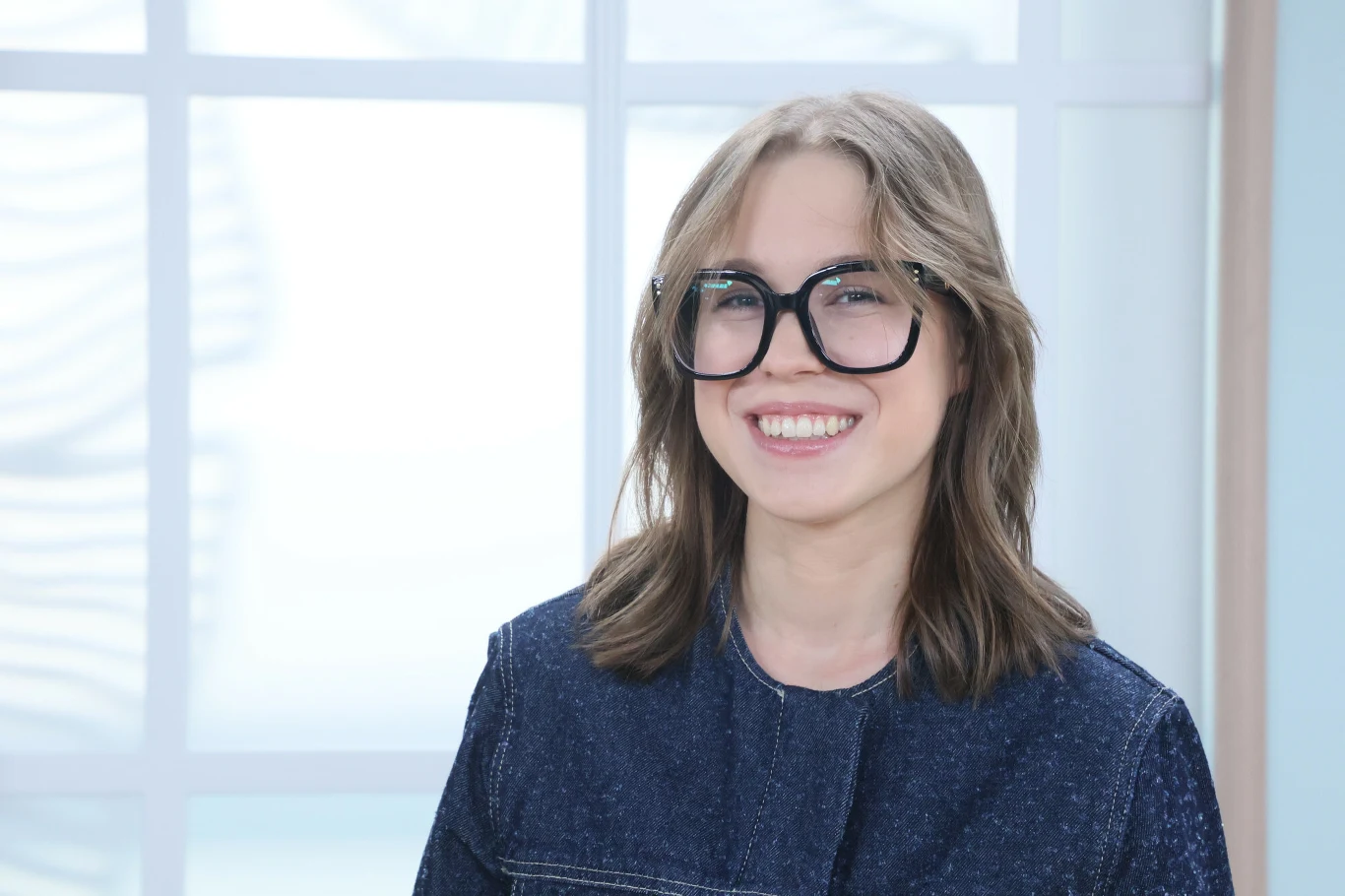 Zoja Skubis Uśmiechnięta młoda kobieta w dużych, czarnych okularach, ubrana w granatową kurtkę na tle jasnego okna.